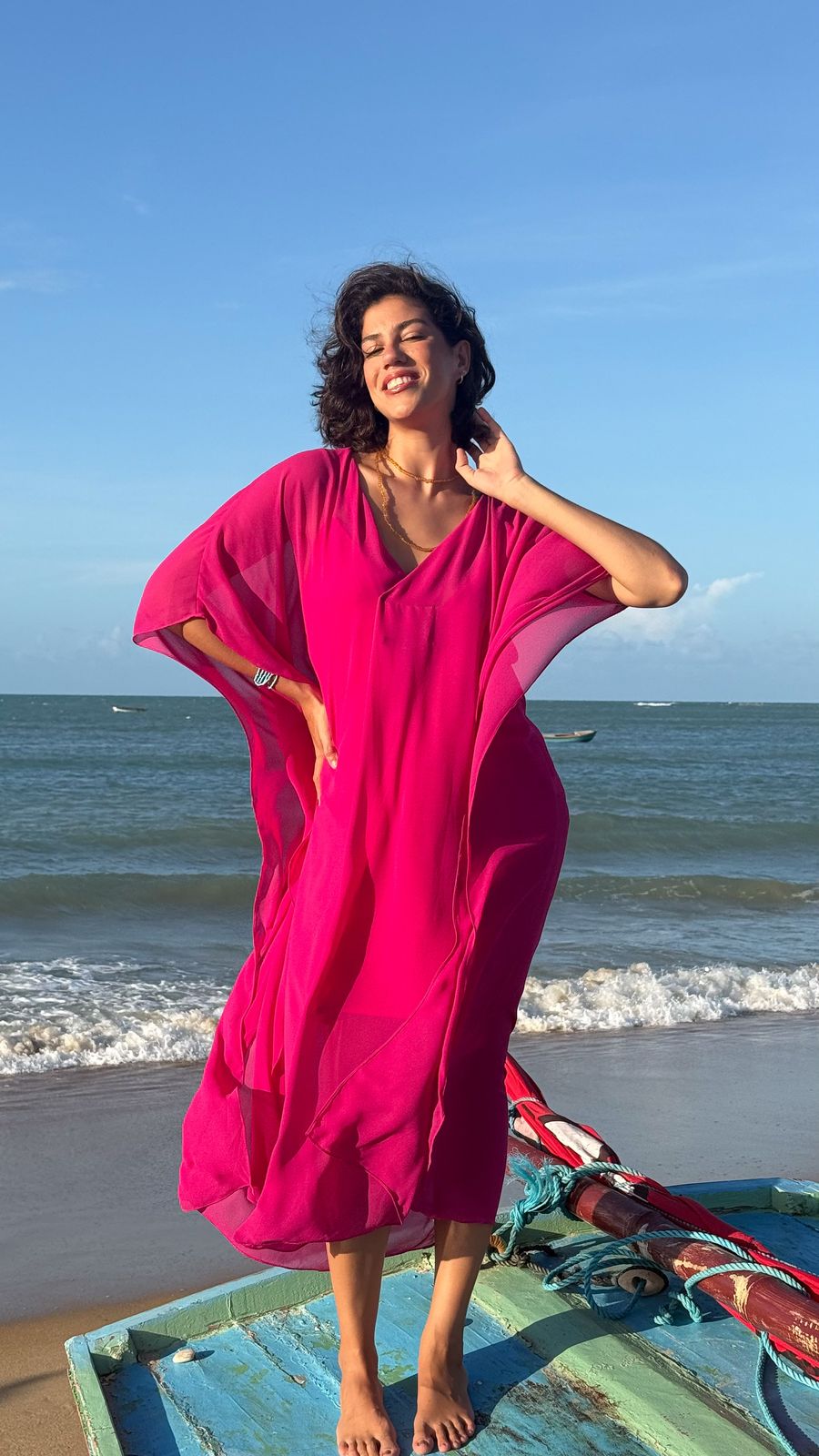 Fernanda Pontes sorrindo em vestido pink, em pé sobre um barco de pesca colorido na praia, luz dourada de fim de tarde