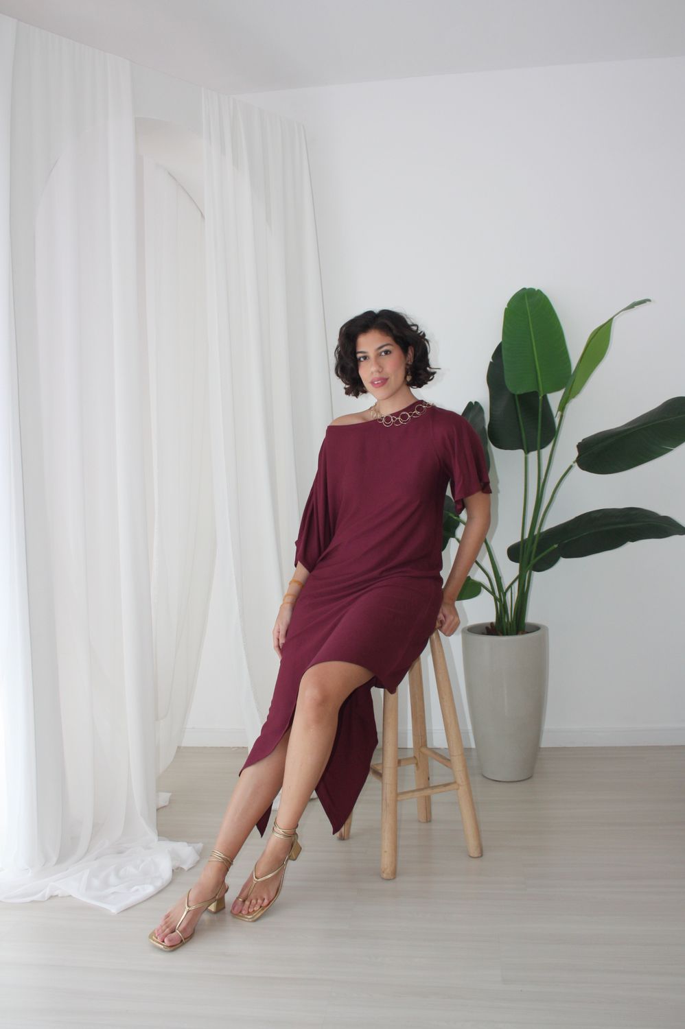 Fernanda Pontes em vestido bordô assimétrico, sentada em banqueta de madeira
