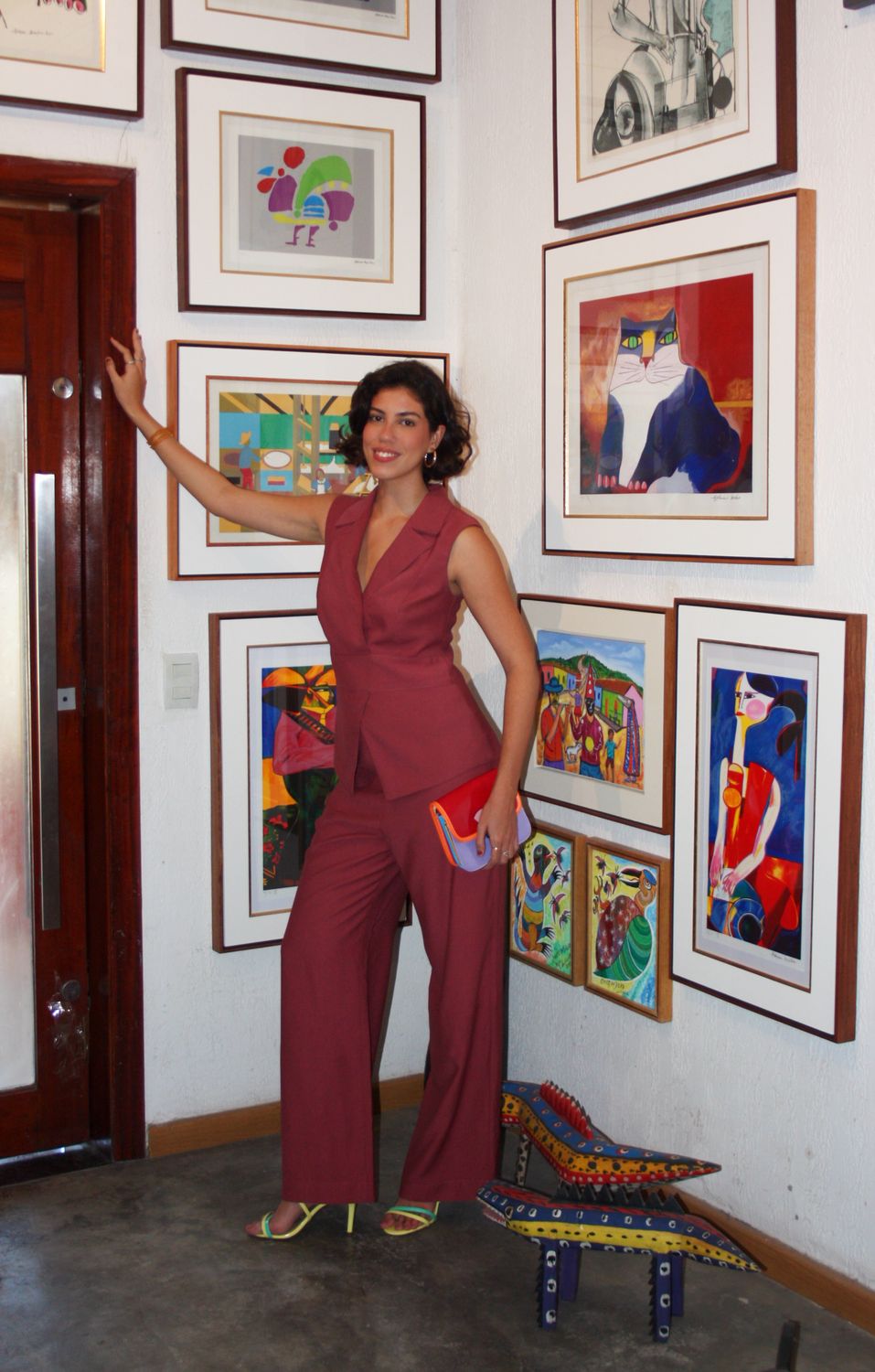 Fernanda Pontes em conjunto vinho, em pé em galeria de arte com quadros coloridos ao fundo