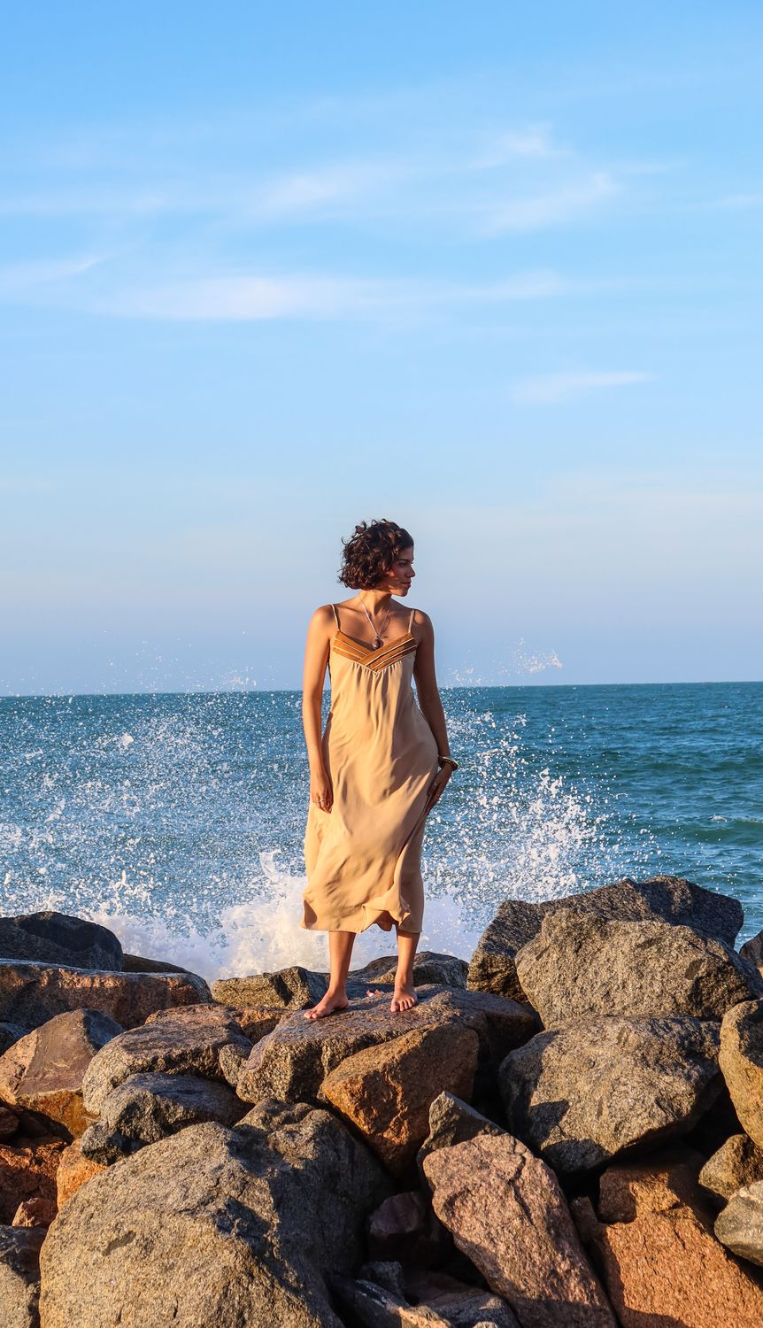 Fernanda Pontes em vestido bege sobre rochas na praia, com onda quebrando ao fundo