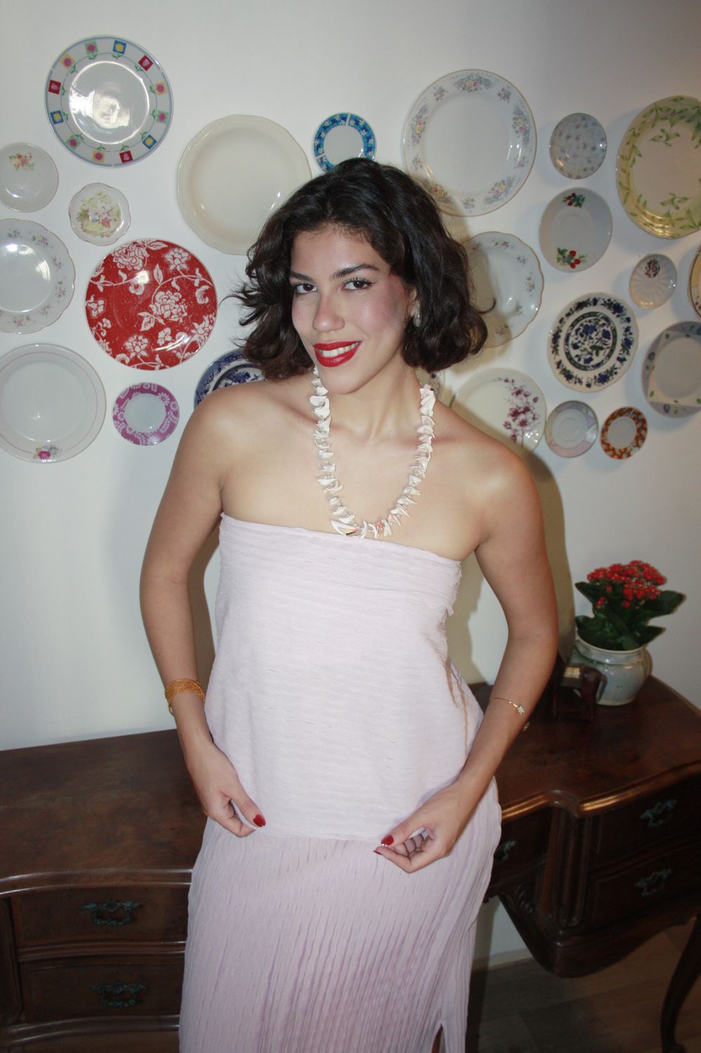 Fernanda Pontes em vestido rosa claro e colar de conchas, em frente a parede decorada com pratos